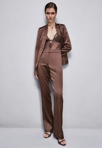 Completo in satin marrone con pantaloni su misura e un blazer abbinato. Sotto è indossato un reggiseno in pizzo nero. Texture liscia, design aderente.