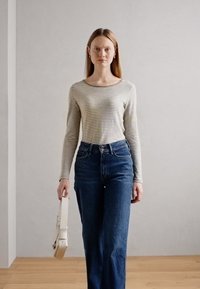Beige és navy csíkos, hosszú ujjú felső, magas derekú kék farmerrel párosítva. A modell krémszínű táskát tart, és fás padlón áll.