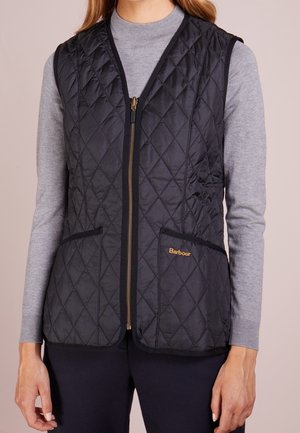 Kobieta ubrana w czarną pikowaną kamizelkę bez rękawów z suwakiem, dwoma przednimi kieszeniami oraz logo "Barbour" na kieszeni, na szarym, długim rękawie.