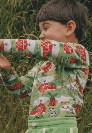 Niño con cabello corto sonriendo, vistiendo una camiseta verde con estampados de graneros rojos, vacas, molinos de viento y tractores, de pie al aire libre cerca de hierba alta.