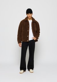 Brun, fluffy, pelsjakke med krage og frontknapper, sammen med en hvit t-skjorte og svart utsvingte jeans.