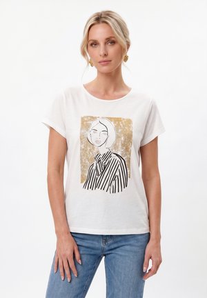 Kvinde iført en hvid T-shirt med et sort og guld kunstnerisk portrættryk, blå jeans og guldbøjler mod en ensfarvet hvid baggrund.