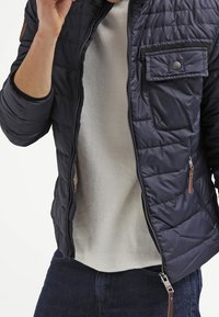 Uomo che indossa una giacca trapuntata blu scuro parzialmente aperta sopra un maglione beige chiaro e jeans scuri, mentre tiene il colletto della giacca.