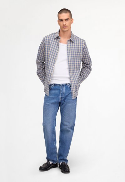 Homme portant une chemise à carreaux bleue sur un débardeur blanc, un jean bleu et des mocassins noirs, debout les mains derrière le dos sur fond blanc.