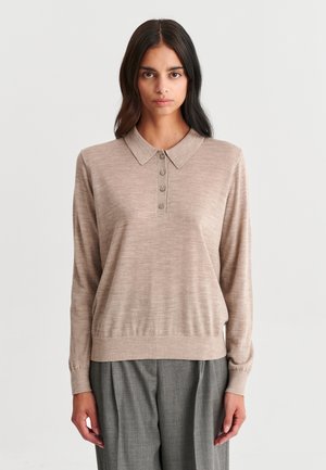 Femme aux longs cheveux foncés, portant un polo beige à manches longues et un pantalon gris, debout devant un fond clair uni.