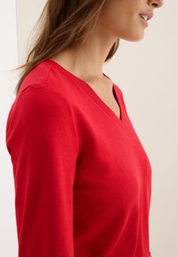 Rotes Langarmoberteil mit V-Ausschnitt, aus glattem Stoff mit taillierter Silhouette. Das Material wirkt weich und bequem.
