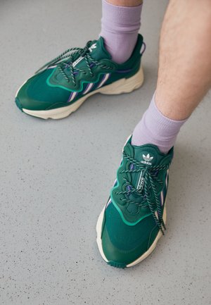Baskets Adidas vertes avec des touches violettes et des lacets rayés portées avec des chaussettes violet clair sur un sol gris moucheté.