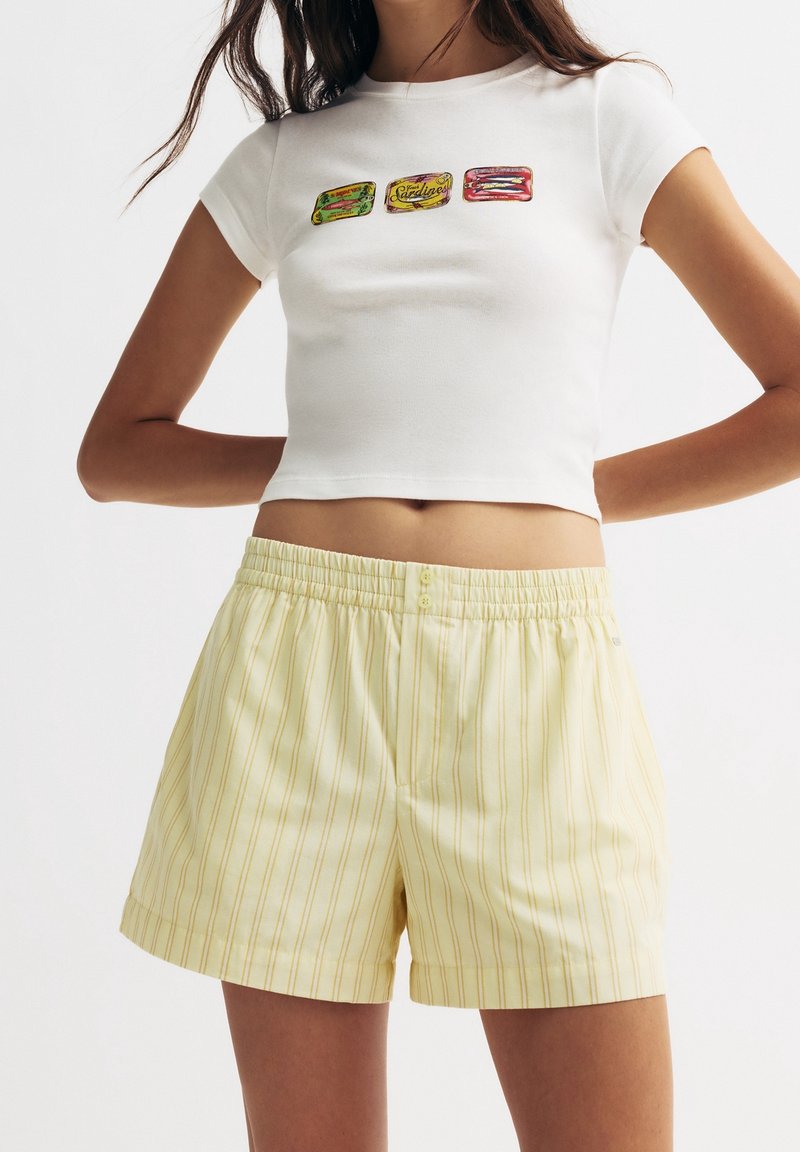 Femme portant un t-shirt court blanc avec des imprimés de boîtes de sardines et un short taille haute jaune clair à rayures verticales.
