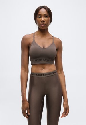 Nő barna Under Armour sportmelltartóban és leggingsben, egy sima világos háttér előtt, szemben állva.