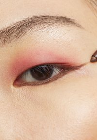 Gros plan sur un œil avec un fard à paupières rose doux se fondant dans la peau, souligné par un eyeliner marron. Les sourcils sont foncés et bien entretenus.