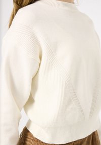 Stickad krämfärgad tröja med ribbat mönster, med rund hals och diagonala stickade detaljer på axlarna. Kort modell.