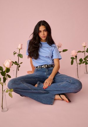 Jonge vrouw met lang donker haar zit met gekruiste benen op een roze vloer, gekleed in een blauw t-shirt en een spijkerbroek, omringd door roze rozen in glazen vazen.