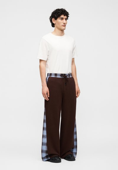 Νεαρός άνδρας στέκεται, φοράει απλό λευκό κοντομάνικο πουκάμισο και καφέ παντελόνι με φαρδιά πόδια με μπλε καρό πλευρικές λωρίδες και μαύρα παπούτσια.