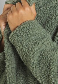 Grön fleece-tyg med en texturerad, fluffig yta. Bilden visar en närbild av materialet och en hand som håller i kanten.
