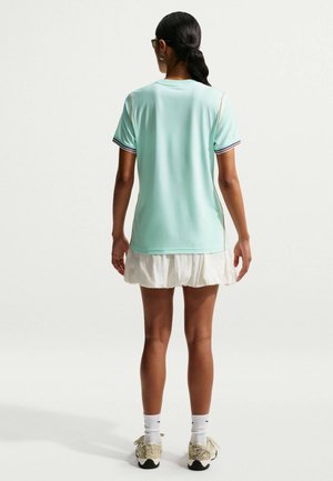 Frau mit zurückgebundenen Haaren, die ein grünes kurzärmeliges Shirt, einen weißen Faltenrock, weiße Socken und helle Turnschuhe trägt, steht mit dem Rücken zur Kamera vor weißem Hintergrund.