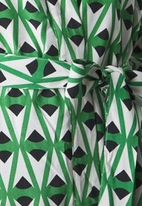 Zelena in bela halja z geometričnim vzorcem črnih oblik. Ima pas, ki je zavezan okoli pasu, in gladko bombažno teksturo.