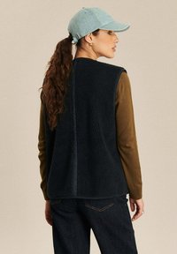 Marine fleece vest met een gestructureerd oppervlak, gedragen over een bruin longsleeve shirt. Een lichtblauwe pet maakt de outfit af. Achteraanzicht.