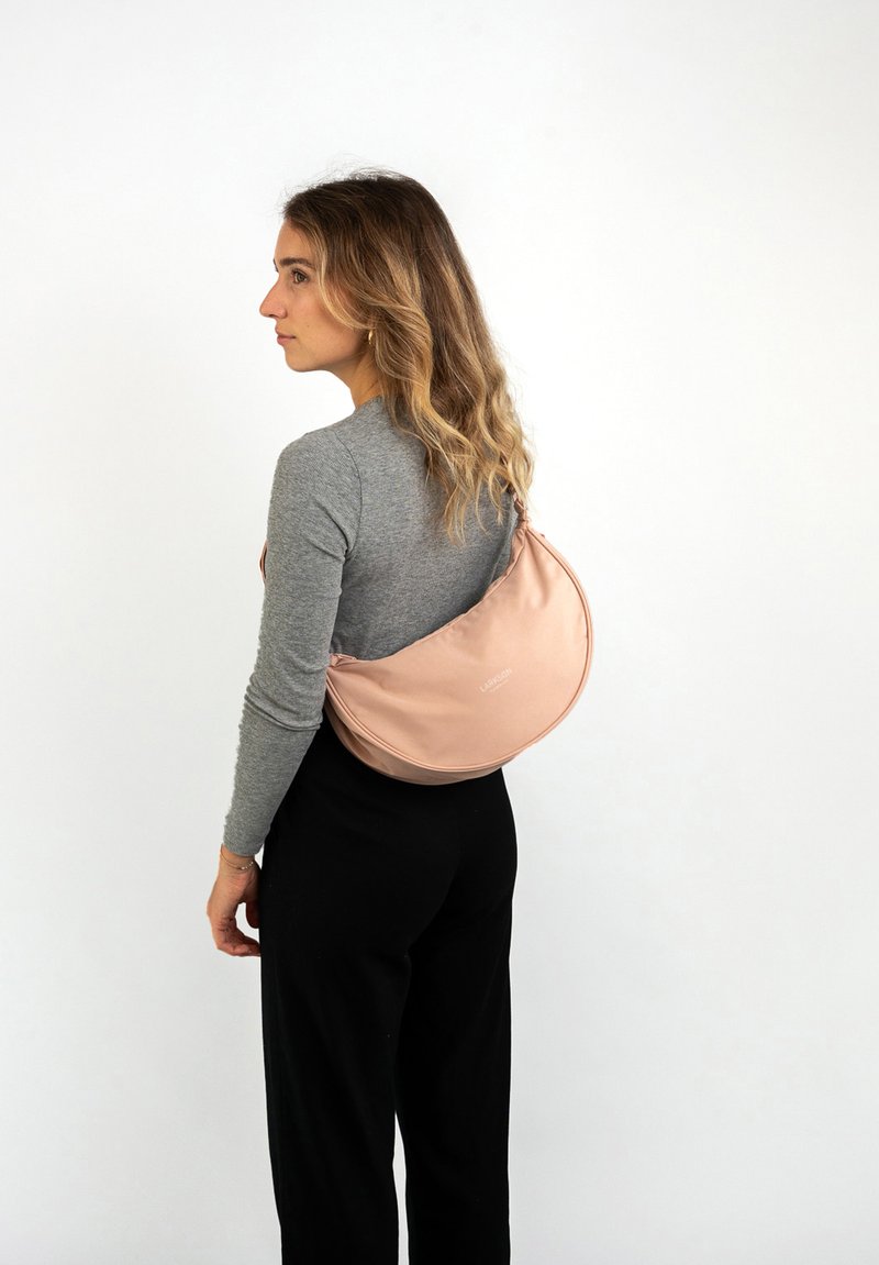 Borsa a tracolla in morbido rosa cipria realizzata in tessuto liscio, con un design curvo e dettagli con logo. Indossata sulla schiena. Include una tracolla regolabile.