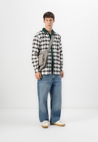 Ung mand iført en grøn skjorte, sort-hvid ternet knap-ops skjorte, brede blå jeans, hvide sneakers og en grå crossover-taske, der står foran en hvid baggrund.