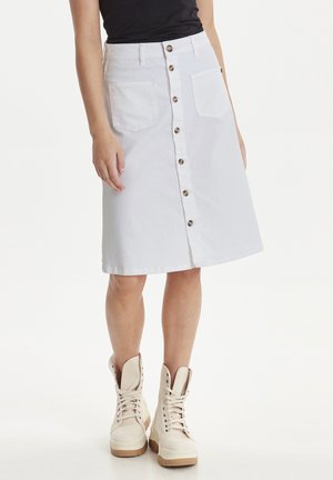 Femme portant une jupe blanche mi-longue boutonnée sur le devant, avec deux poches avant et des bottines beiges à lacets.