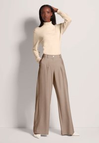 Beige coltrui gecombineerd met hoogstaande, brede taupe broeken met voorplooien en een enkele knoopsluiting. Effen witte schoenen.