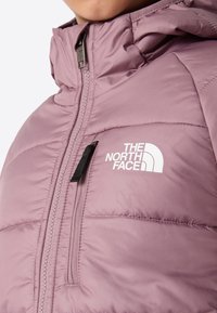 Zbliżenie na różową, pikowaną kurtkę, z pionową kieszenią na zamek błyskawiczny na piersi i białym logo "The North Face" z przodu.