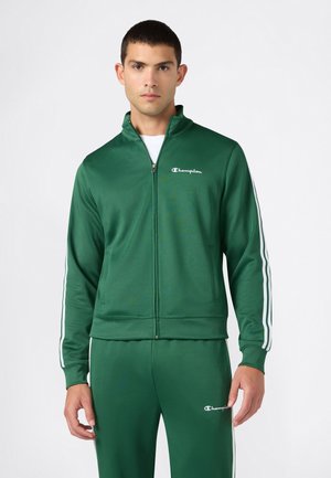Mann trägt einen grünen Champion-Trainingsanzug mit weißen Streifen an Ärmeln und Hose, steht vor einem schlichten weißen Hintergrund.