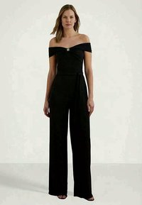 Macacão preto sem mangas com corpete ajustado, calças de perna larga e um detalhe em nó na cintura, apresentando um acabamento prateado.