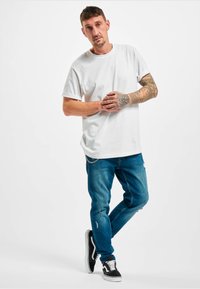 Camiseta blanca de algodón, con cuello redondo, combinada con jeans azul desgastados. Zapatillas negras con acentos blancos y un diseño de perfil bajo.