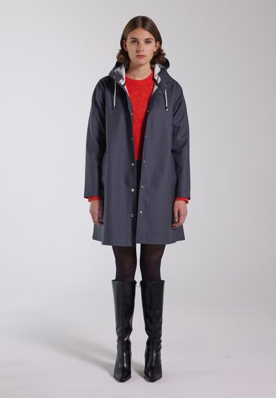 Grå regnfrakke med hætte, med en hvid inderskrald og trykknaplukning. Båret over en rød sweater og parret med sorte knæhøje støvler.