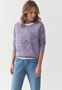 Felpa viola con motivo floreale ricamato al centro, maniche lunghe, scollatura rotonda, indossata sopra una camicia bianca, abbinata a jeans blu.