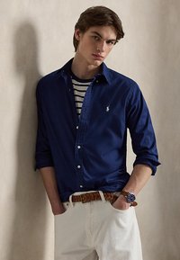 Fiatal férfi, aki sötétkék, gombos inget visel csíkos póló fölött, fehér nadrágot, barna fonott övet és karórát, és egy fal mellett áll.