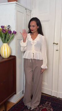 Femme en blouse blanche à volants et pantalon beige faisant un signe de la main, debout à côté d'une armoire blanche avec un vase rayé jaune contenant des fleurs violettes.