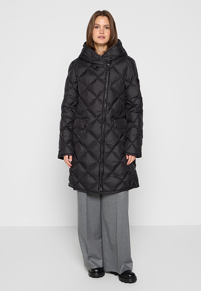 Donna che indossa un lungo cappotto imbottito nera con cappuccio, pantaloni grigi a gamba larga e scarpe nere, in piedi su uno sfondo bianco semplice.