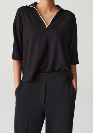 Woman wearing a loose black top with white-trimmed collar and black high-waisted pants, standing with hands in pockets.