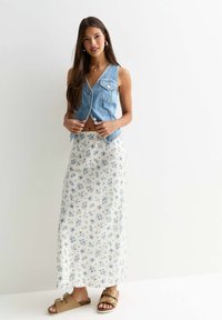 Jeansväst med knappar och sidofickor, i kombination med en blommig maxikjol i vitt och blått. Bruna sandaler med spännen fulländar looken.