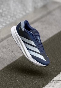 Blue and white Adidas running shoe in mid-air above asphalt with textured concrete edge in background.