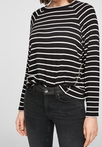 Schwarz-weißer gestreifter Langarmoberteil mit Rundhalsausschnitt; kombiniert mit schwarzen Jeans mit einem Reißverschluss-Detail an der Seite.