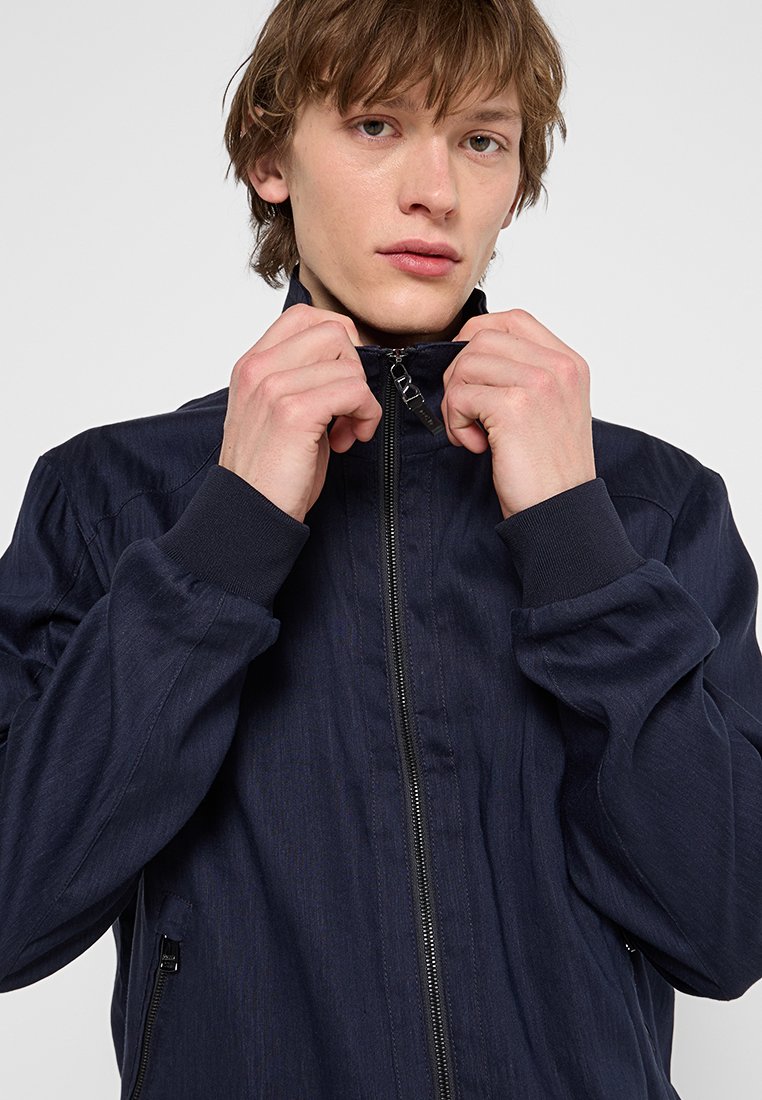 Marineblaue Zip-Jacke mit hohem Kragen, gerippten Bündchen und seitlichen Taschen. Glatte Textur mit dezenten vertikalen Mustern. Die Hände des Models passen den Kragen an.