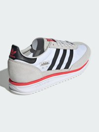 Witte sneaker met zwarte strepen, grijze suède accenten en rode details op de zool, met een gevoerde kraag en logo.