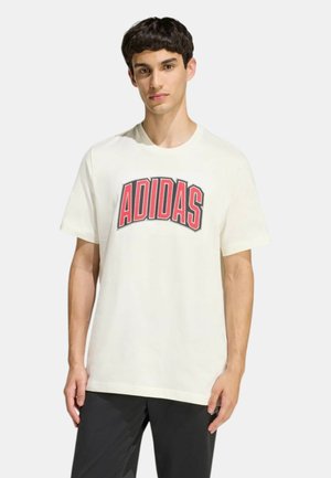 Giovane uomo con maglietta bianca con scritta rossa "ADIDAS" e pantaloni neri, in piedi contro uno sfondo chiaro e uniforme.