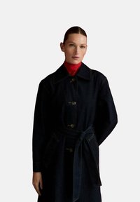 Manteau trench en denim foncé avec une taille ceinturée, un large col et une fermeture à boutons. Présente des coutures contrastées et une texture lisse.
