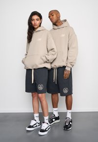 Felpe beige e pantaloni corti grigi scuri con dettagli del logo. I modelli indossano sneakers, mostrando un'estetica streetwear casual.