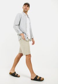 Lys grå stribet skjorte med knapper, hvid t-shirt, beige shorts og mørke sandaler med to spænder, præsenteret i en afslappet gangposition.