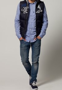 Mörkblå puffväst med dragkedja, broderade vita och blå logotyper på framsidan, bärs över en ljusblå skjorta med knappar och blå jeans.