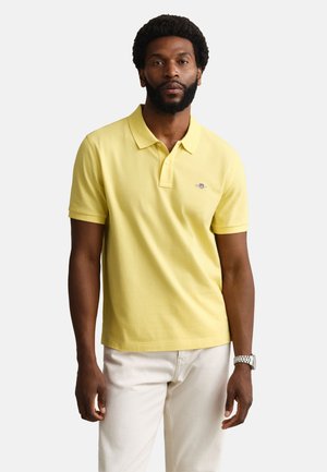 Hombre con barba que lleva una camisa polo amarilla de manga corta y pantalones blancos, de pie frente a un fondo claro y liso, reloj en la muñeca izquierda.