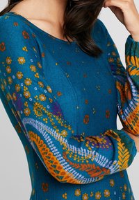 Femme portant une robe bleu sarcelle avec des motifs floraux et à motifs orange, violet et jaune sur les manches et le corsage.