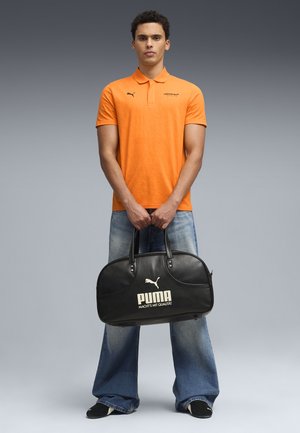 Hombre con polo naranja y jeans anchos sosteniendo un bolso de viaje negro Puma contra un fondo gris.