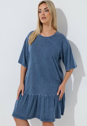 Blondynka ubrana w luźną, niebieską sukienkę z krótkim rękawem i marszczonym dołem, stojąca na tle gładkiej białej ściany.
