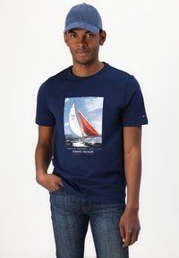 Mørkeblå bomulls t-skjorte med et fargerikt seilbåt grafikk med røde og hvite seil. Kombinert med mørke jeans og en lyseblå caps.